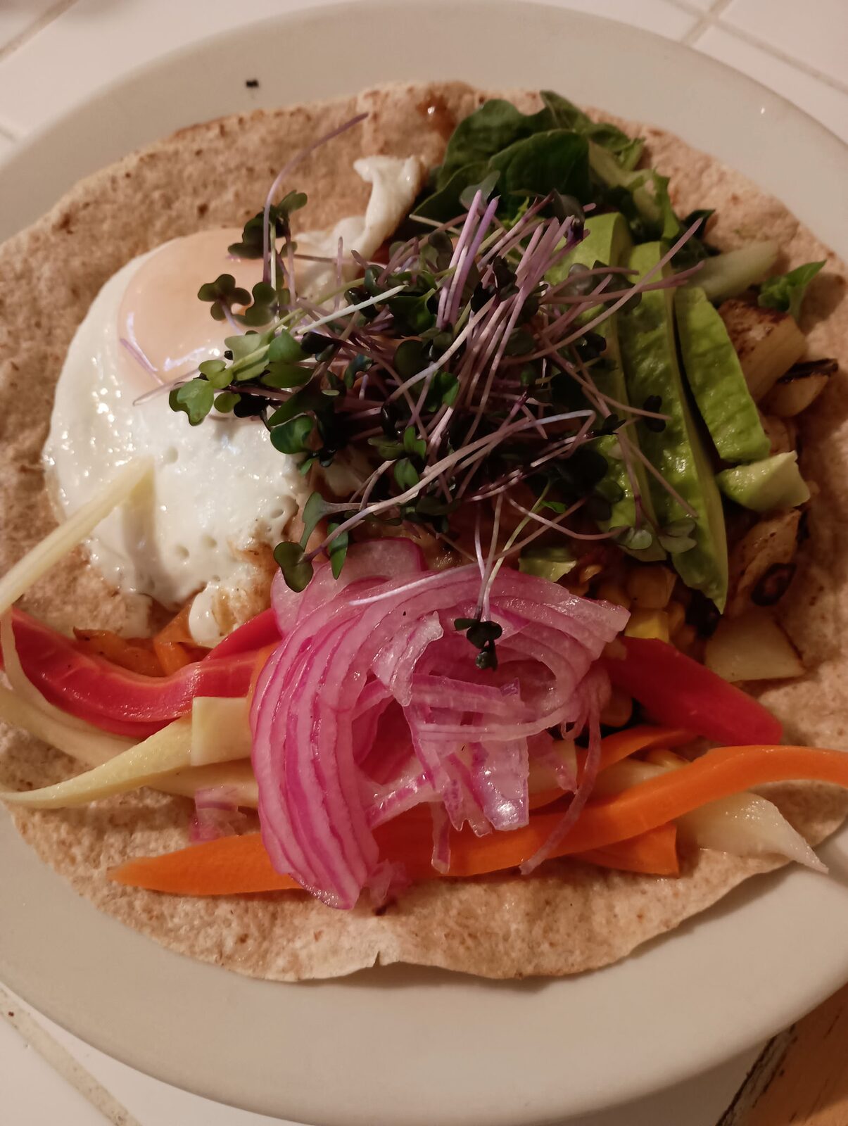A whole-wheat tortilla topped with a sunny-side-up egg, pickled red onions, vegetables, and microgreens.