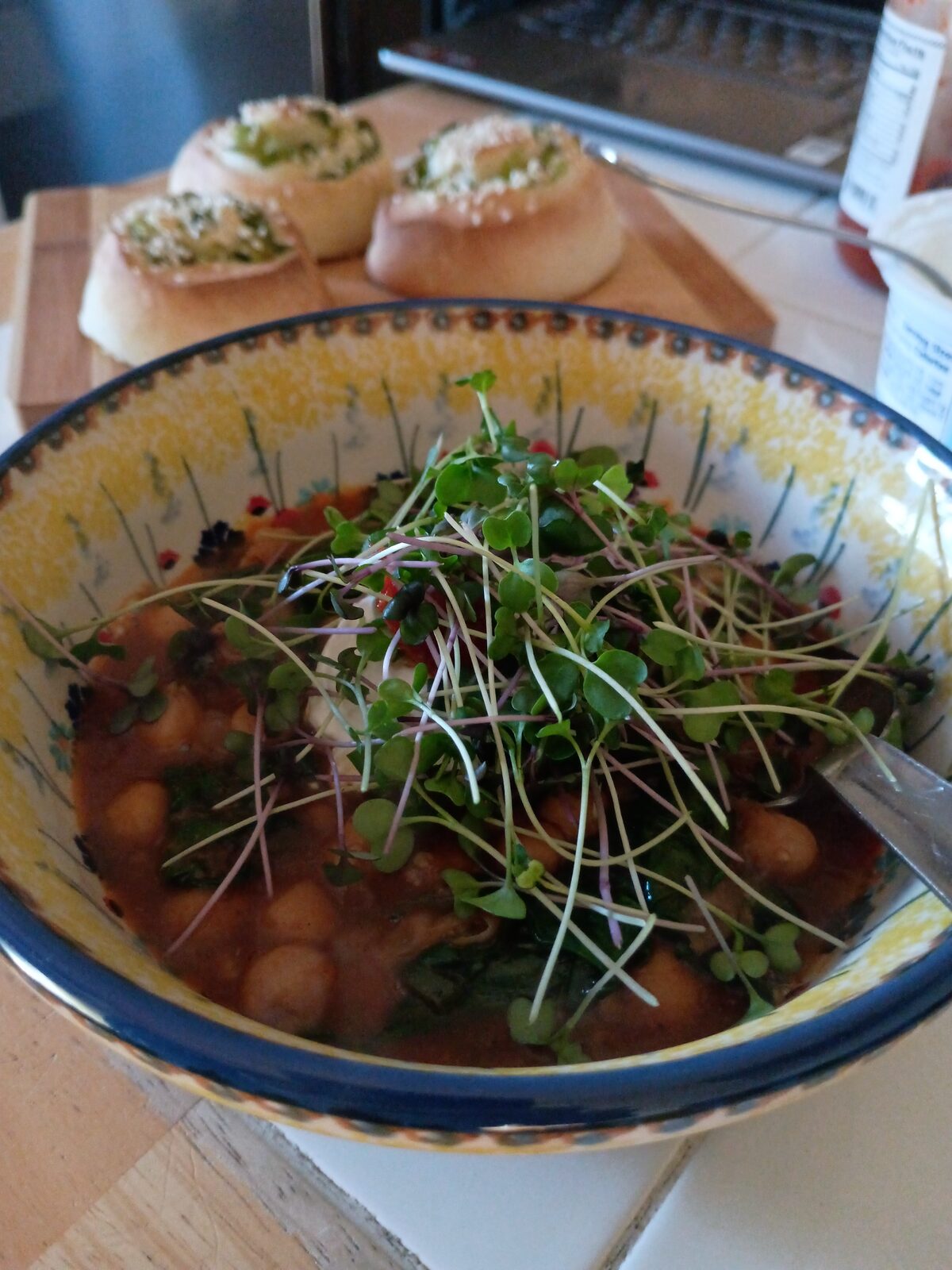 A bowl of stew with chickpeas, topped with a generous tangle of fresh microgreens, served alongside herbed bread rolls.