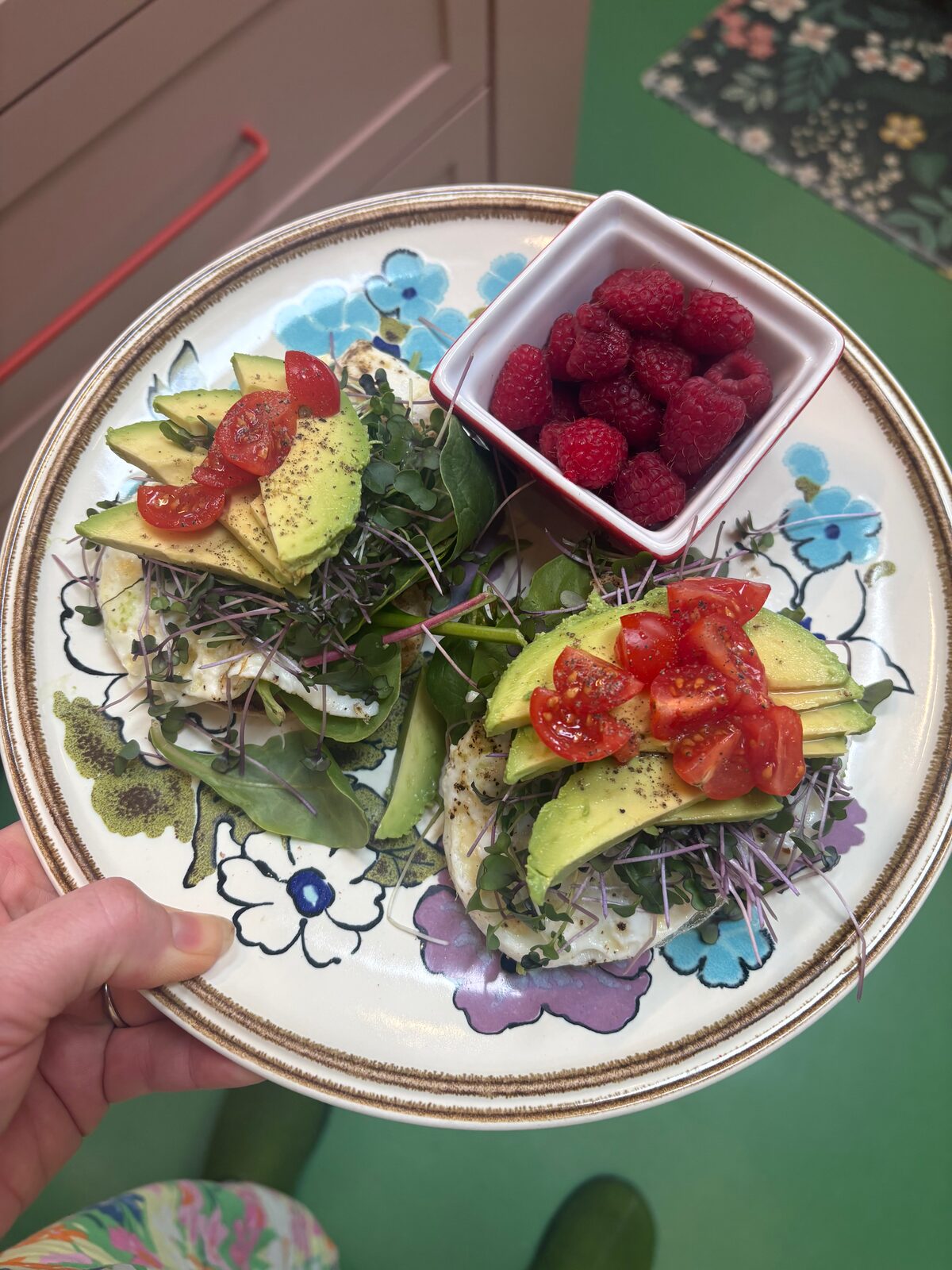 A floral plate of avocado toast with sliced tomato, microgreens, and a small dish of fresh raspberries.